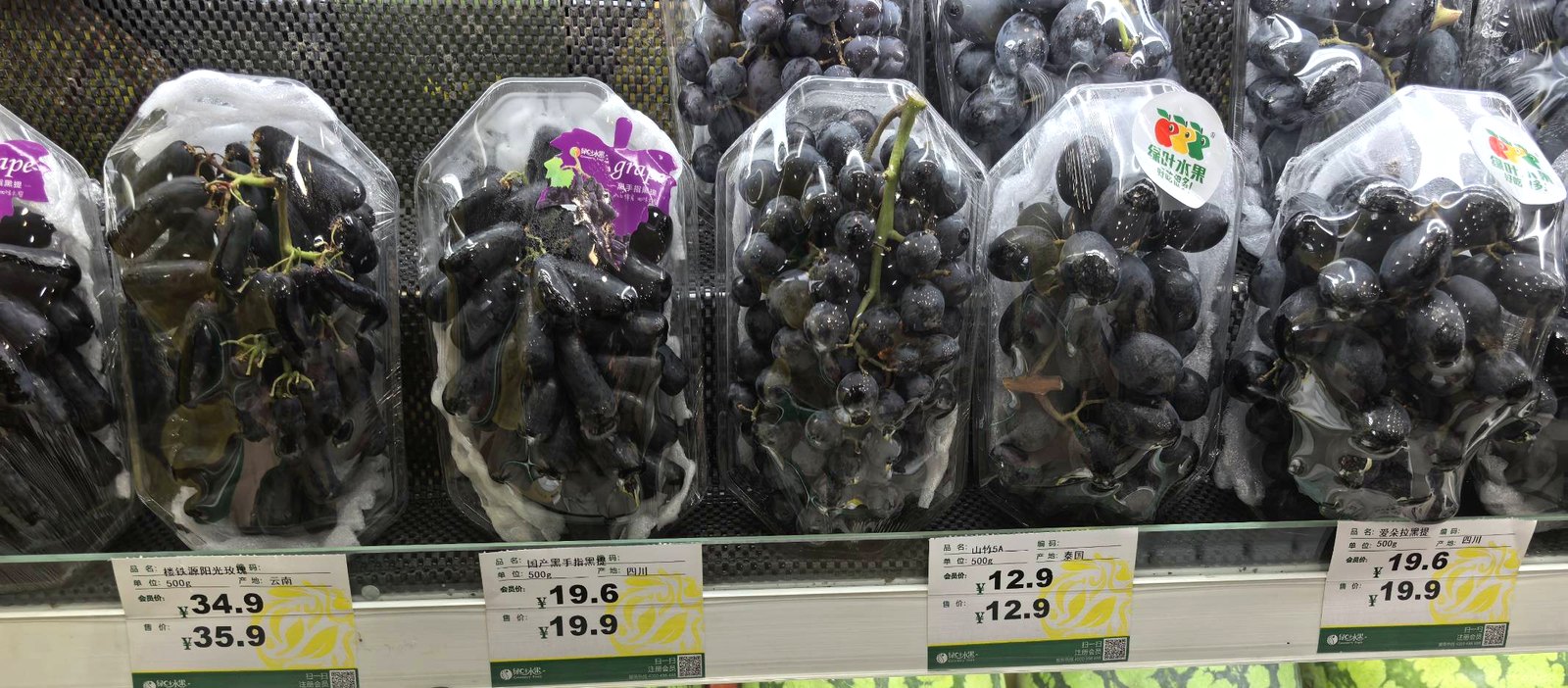 绿叶水果门店葡萄区货架，同一排陈列着阳光玫瑰、黑手指黑提、爱朵拉黑提等不同品种，价签从 19.9 到 35.9 不等